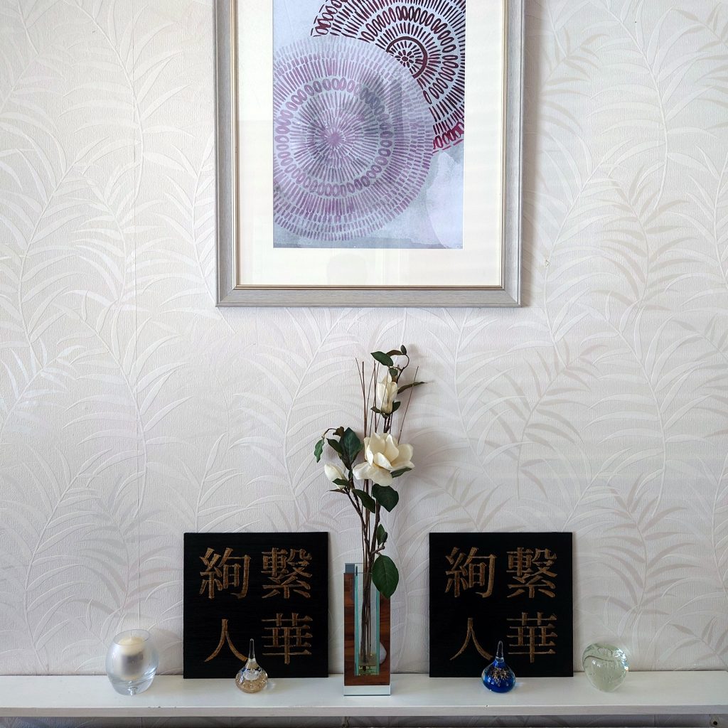 two identical black and gold acrylic Japanese kanji paintings on white shelf beside white flowers, with small glass ornaments