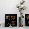 Ayatsuna Hitoka 絢繋 | 人華 BY NIGHT Acrylic Painting - Photo of a black and gold acrylic Japanese kanji painting on white shelf beside white flowers, with small glass ornaments