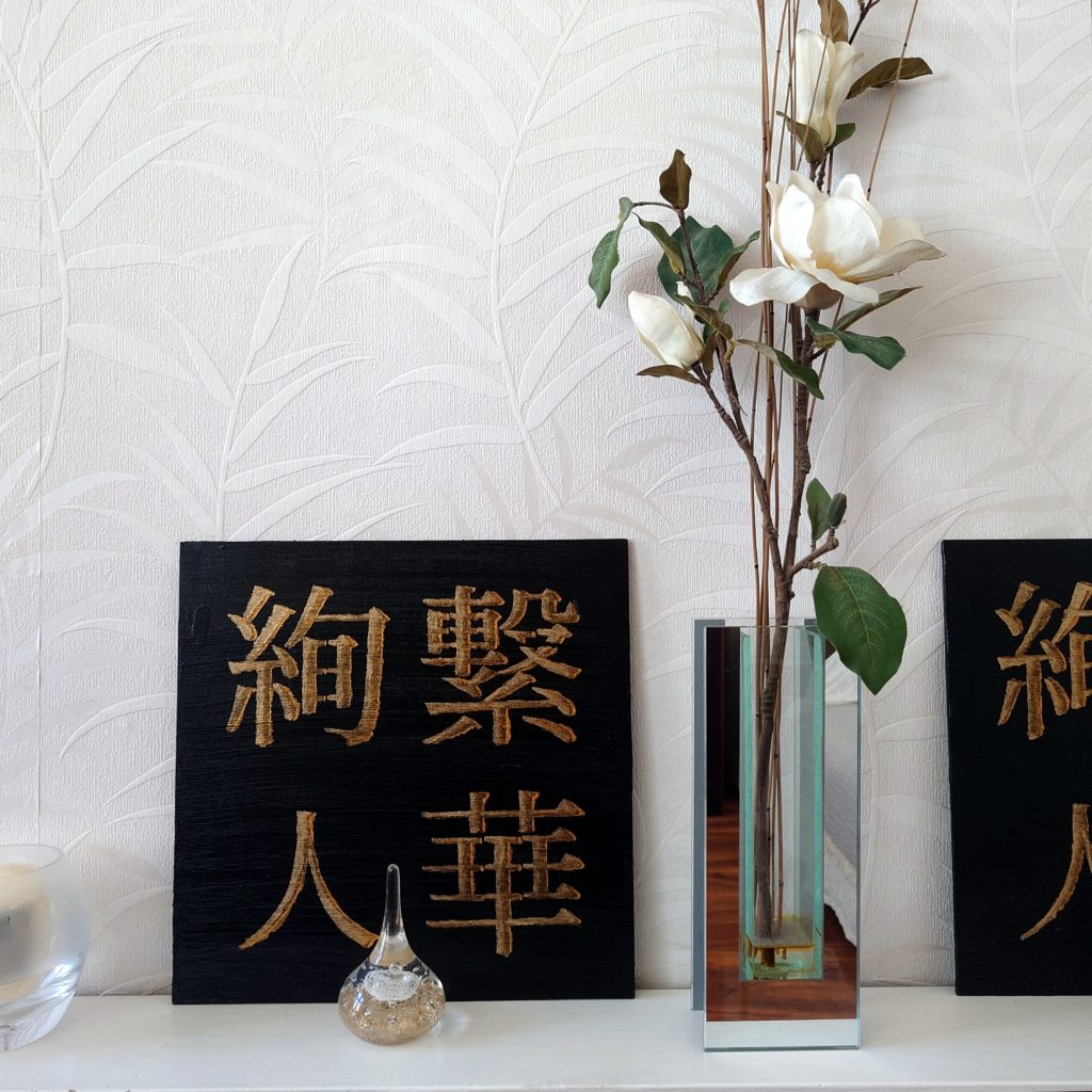 black and gold acrylic Japanese kanji painting on white shelf beside white flowers, with small glass ornaments
