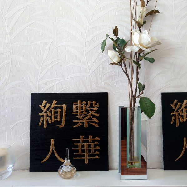 Ayatsuna Hitoka 絢繋 | 人華 BY NIGHT Acrylic Painting - Photo of a black and gold acrylic Japanese kanji painting on white shelf beside white flowers, with small glass ornaments