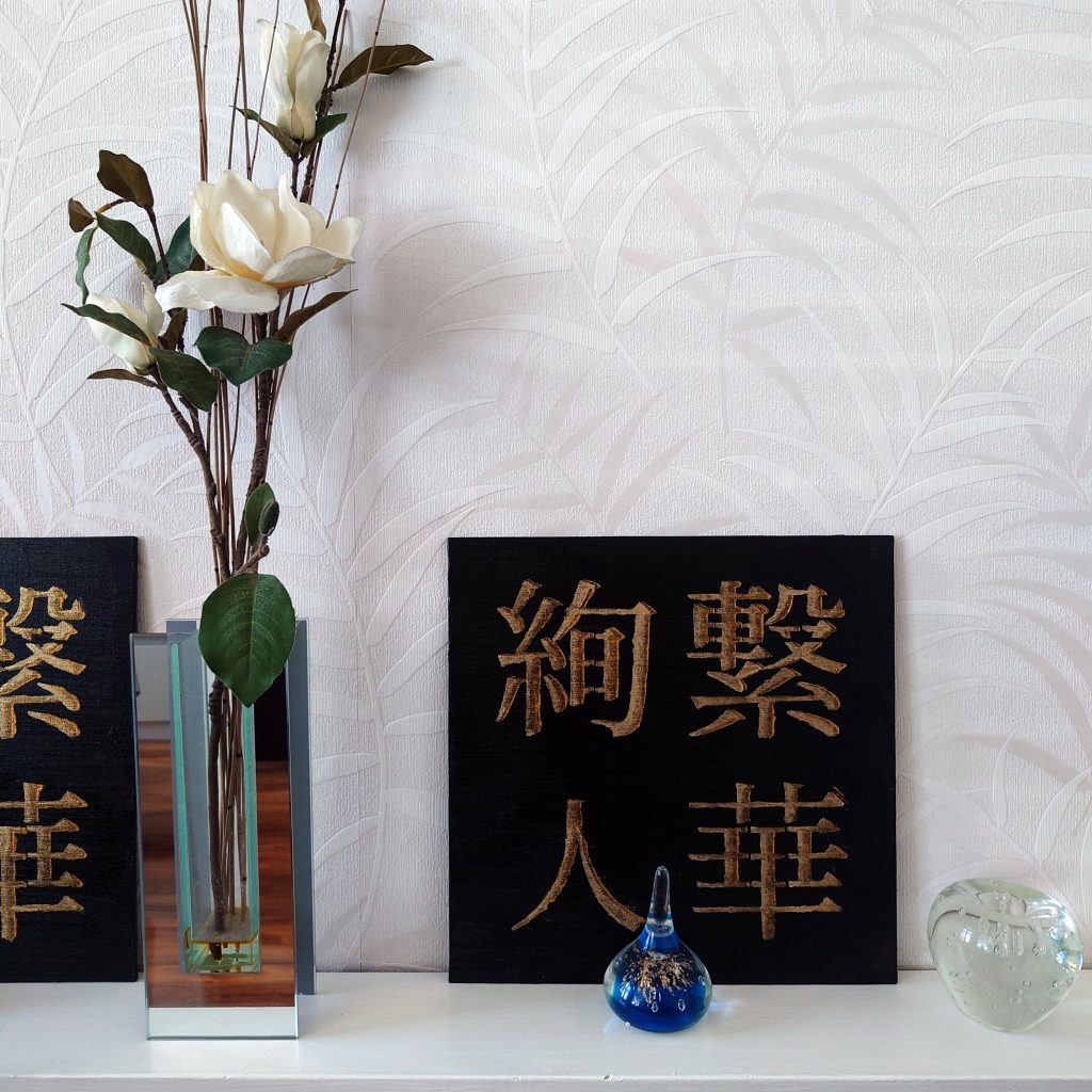 black and gold acrylic Japanese kanji paintings on white shelf beside white flowers, with small glass ornaments