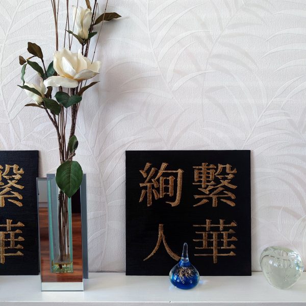 Ayatsuna Hitoka 絢繋 | 人華 BY NIGHT Acrylic Painting - black and gold acrylic Japanese kanji paintings on white shelf beside white flowers, with small glass ornaments